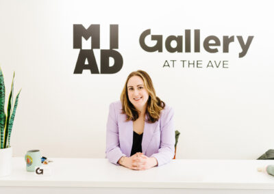 A person in a purple jacket sits behind a white desk. MIAD Gallery at the Ave logo is on the wall behind them.