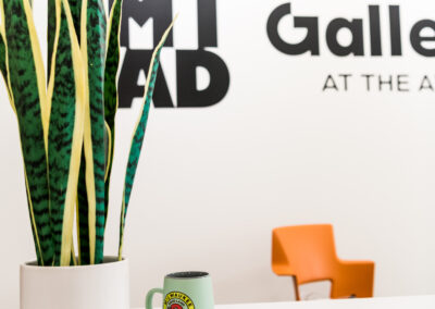Empty white desk with plant and mug in foreground. In the background is the MIAD Gallery at the Ave logo.