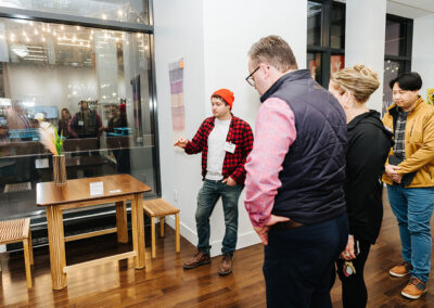 Group of people stand in Gallery at the Ave. One person is speaking and pointing to a wooden table.