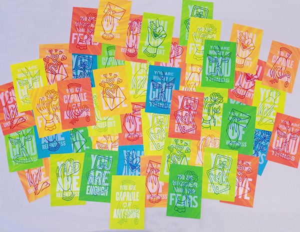 A gallery wall filled with colorful screenprinted postcards with positive affirmations on them.